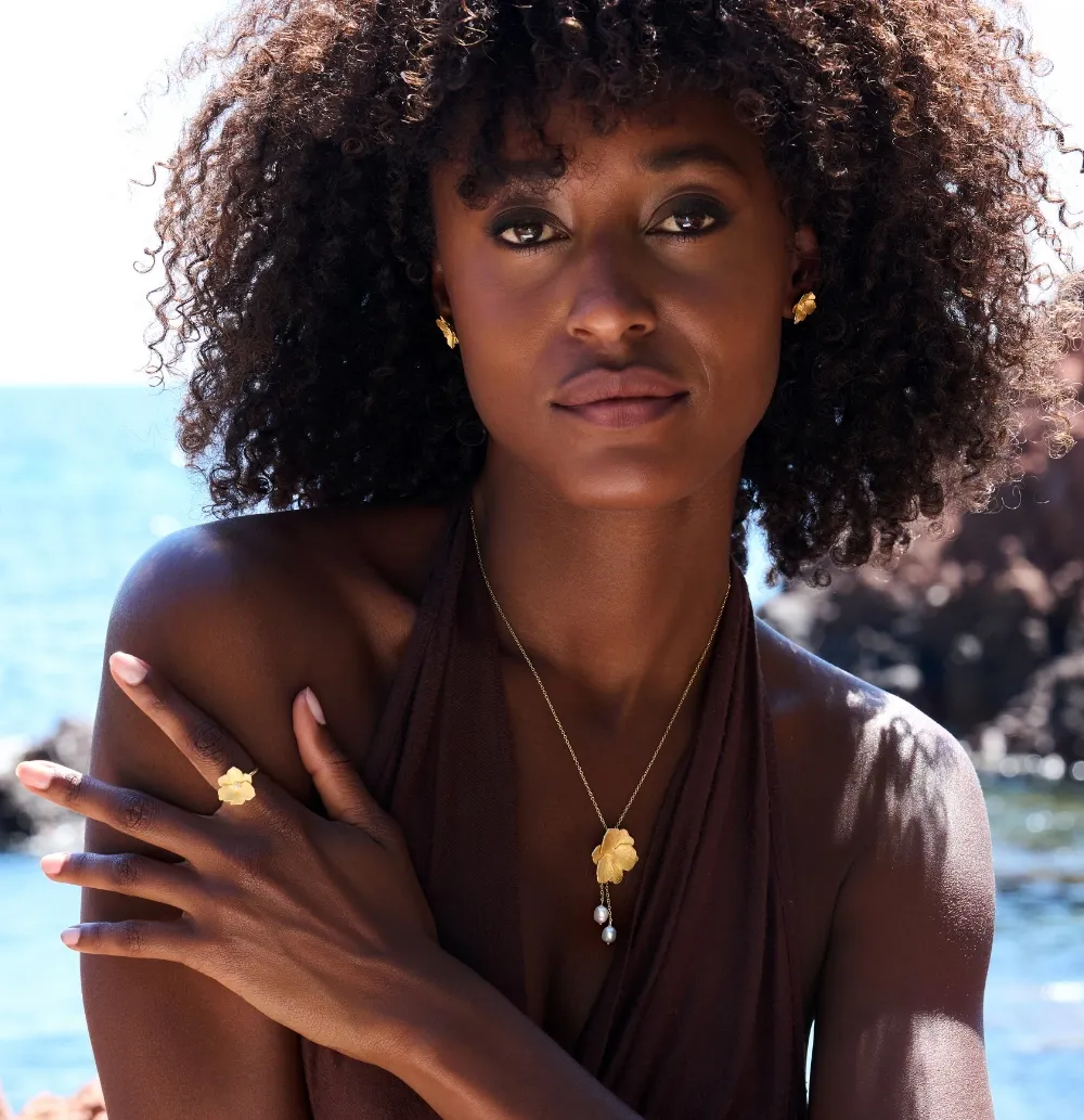 Elegant woman with curly hair wearing gold jewelry, necklace, ring, and earrings, outdoors.