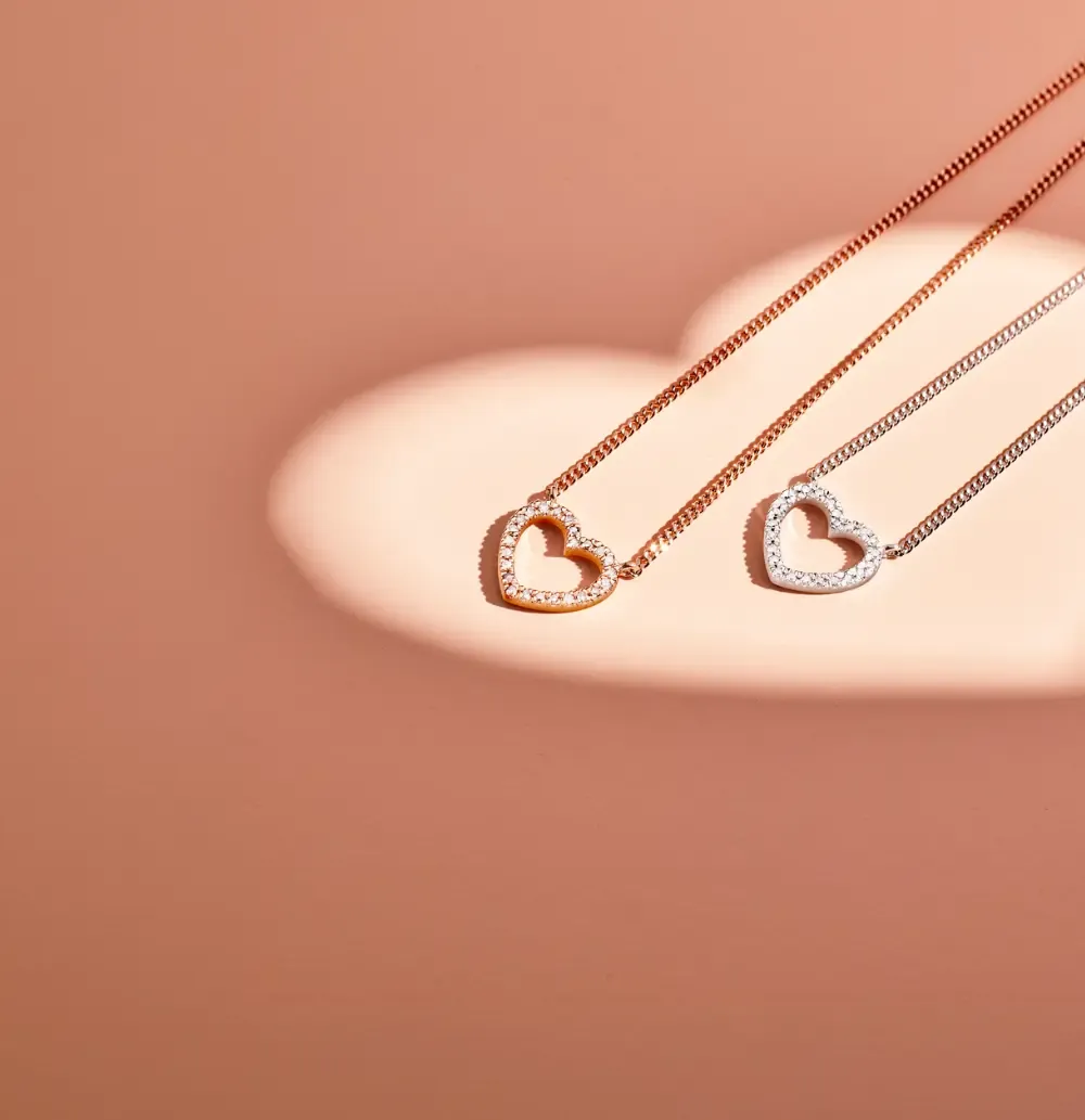 Two sparkling heart pendant necklaces, one rose gold and one silver, on a pink surface.