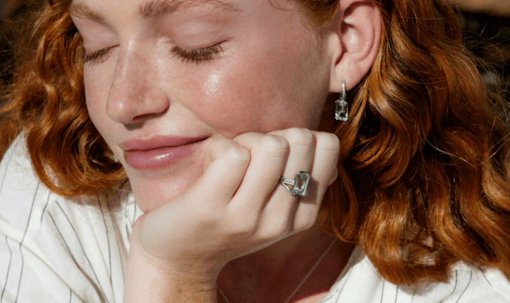 Redhead woman with closed eyes, wearing aquamarine ring and earrings, basking in sunlight.